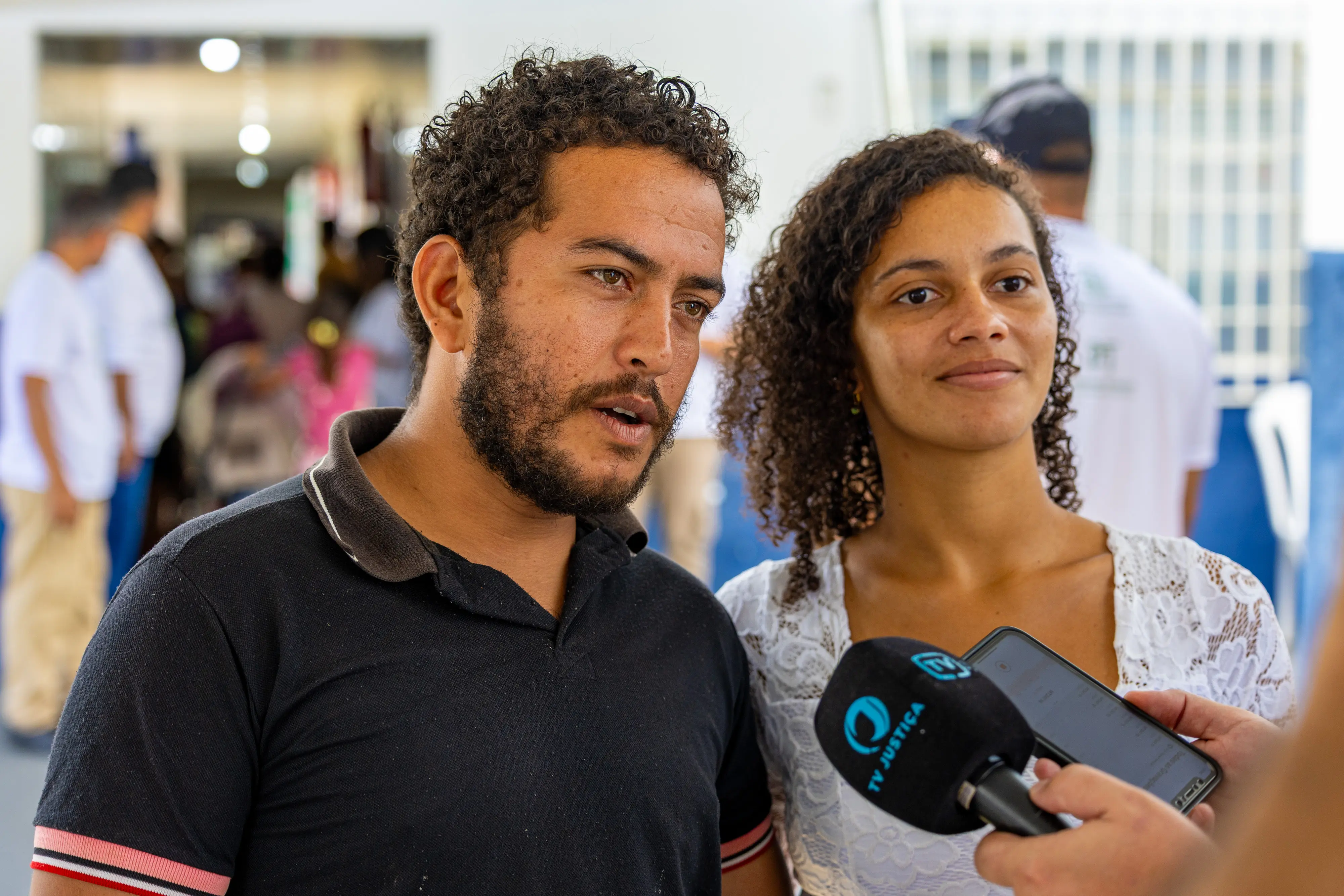 Em primeiro plano, o casal concede entrevista a uma equipe de comunicação. Um microfone com identificação institucional é direcionado a eles, enquanto o homem fala e a mulher observa. Ao fundo, outras pessoas circulam, indicando movimento e atendimento coletivo.