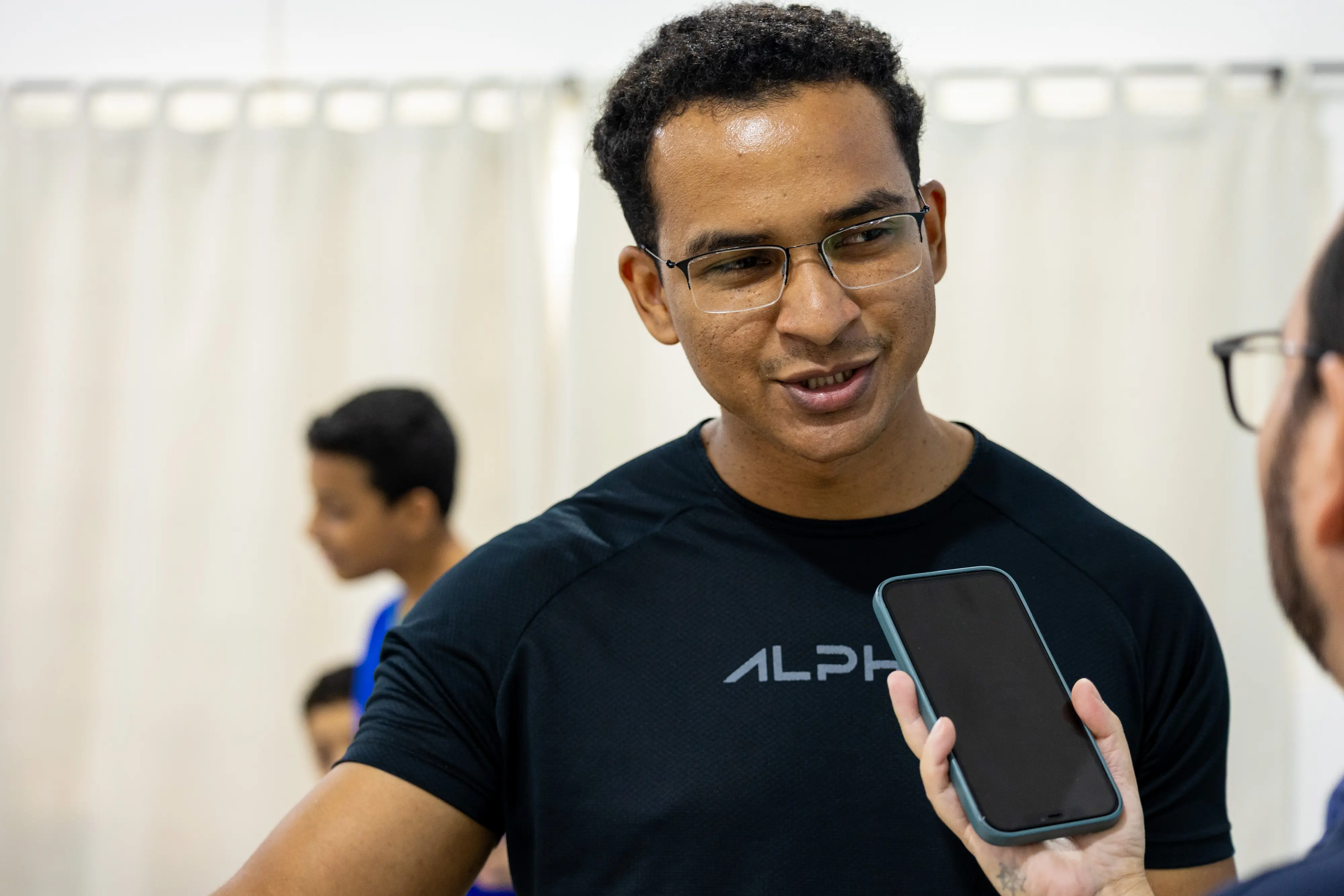 Homem jovem de óculos e camiseta preta fala sorrindo enquanto é entrevistado. Uma mão segura um celular à frente dele para gravar. Ao fundo, vultos de estudantes e uma cortina clara.
