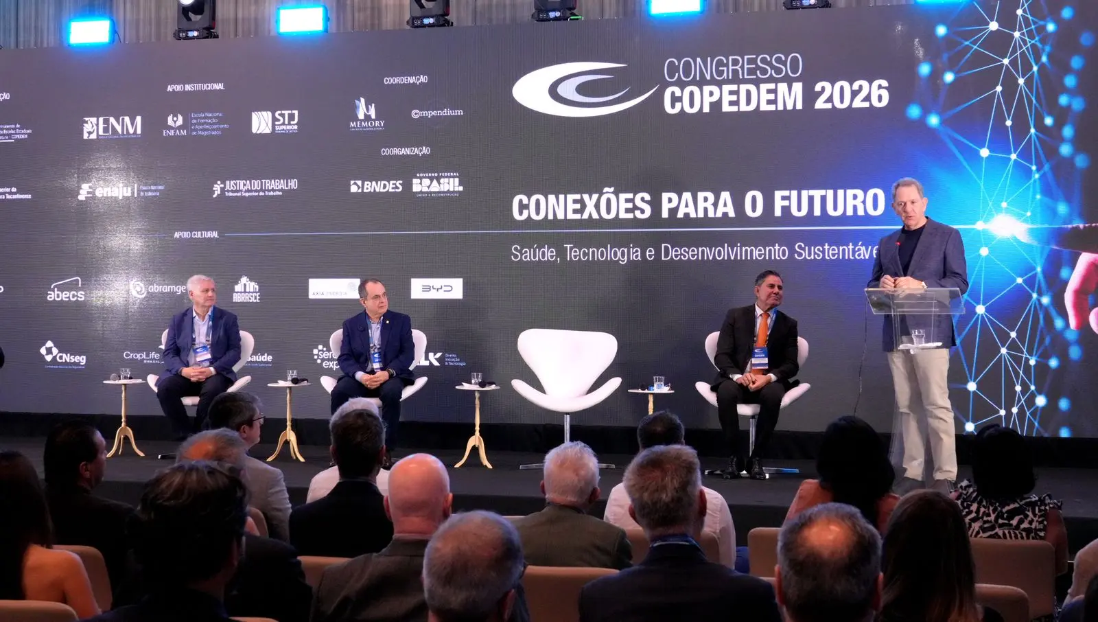 Foto colorida. Homem fala ao microfone, da tribuna. Ele veste terno e camisa escuros, além de calça beje. Ao lado dele, três outros homens sentam em cadeiras alinhadas uma ao lado da outra. 