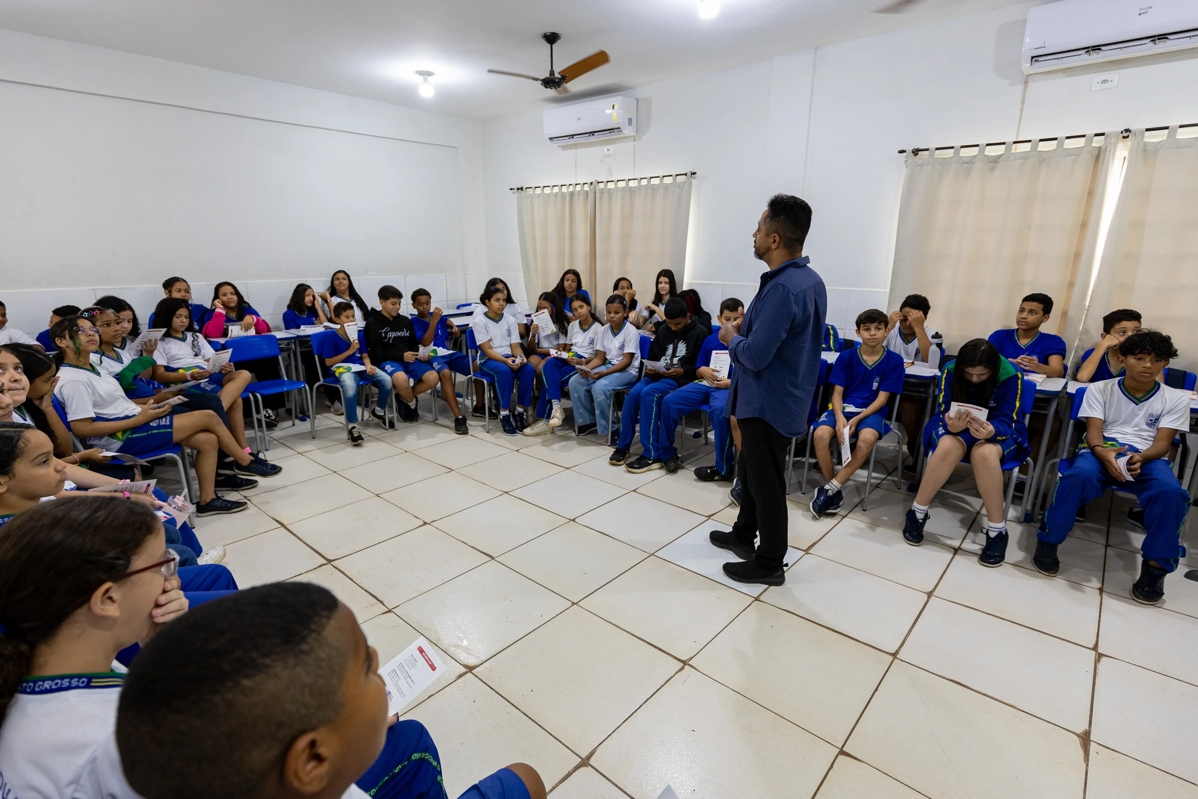 Um homem de pé fala para um grupo de estudantes sentados em círculo em uma sala de aula. Os jovens vestem uniformes azuis e brancos e seguram folhetos. O ambiente é iluminado e tem paredes claras.