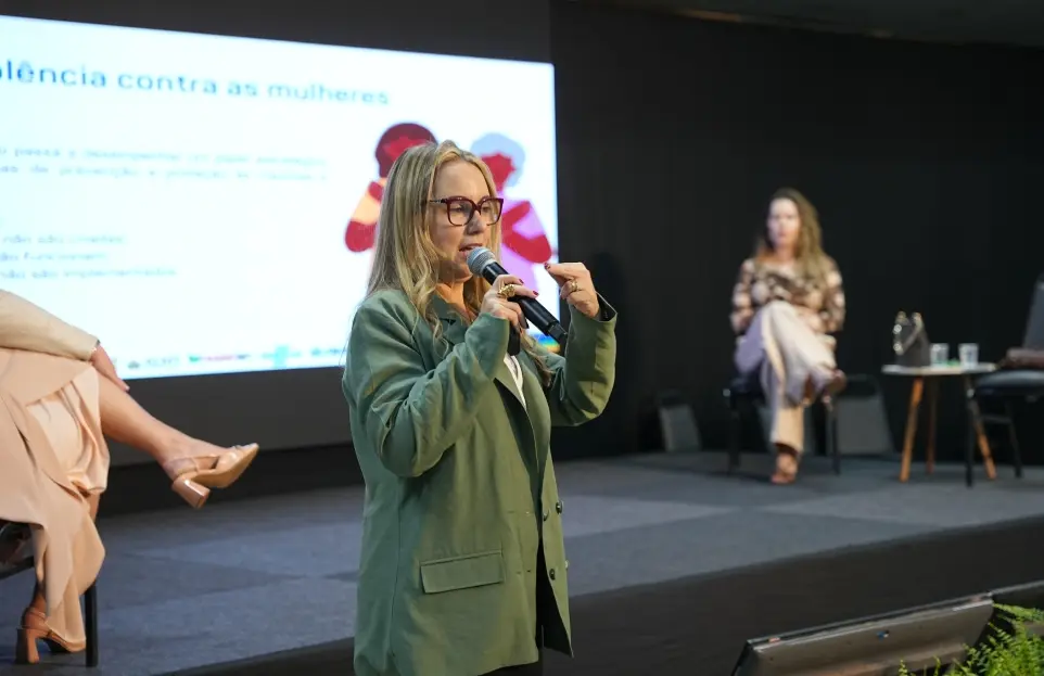 Foto horizontal que mostra a juíza Ana Graziela Vaz de Campos em pé, da cintura pra cima, falando ao microfone, na parte debaixo do placo de um auditório. Ela é uma mulher branca, loira, usando óculos de grau, blusa branca e blazer verde. 