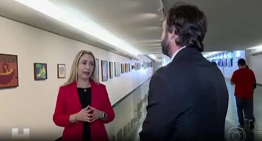 Mulher de blazer vermelho conversa com homem de terno em corredor amplo, com quadros alinhados na parede. Ao fundo, pessoa caminha. A cena indica entrevista em ambiente institucional.